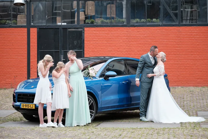 Marleen en Eric met de bruidsmeisjes bij een blauwe auto voor rode baksteen