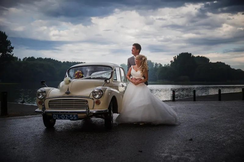 Bruidspaar bij een crèmekleurige Morris Minor aan het water onder een bewolkte hemel
