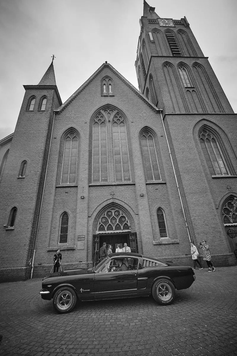 Zwart-wit: zwarte Ford Mustang voor een bakstenen kerk, het bruidspaar in de auto, gasten op de trap