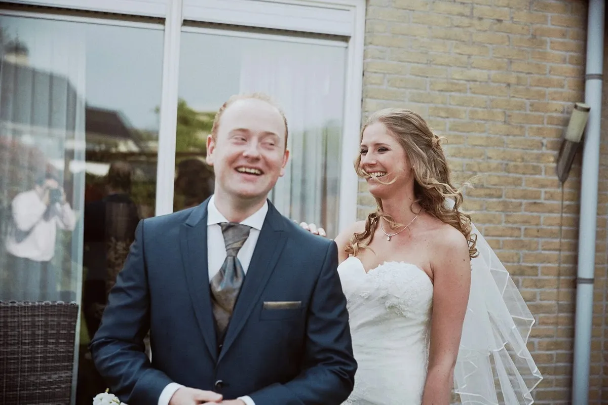 Het bruidspaar lacht samen buiten tegen een bakstenen muur, vlak na de first look