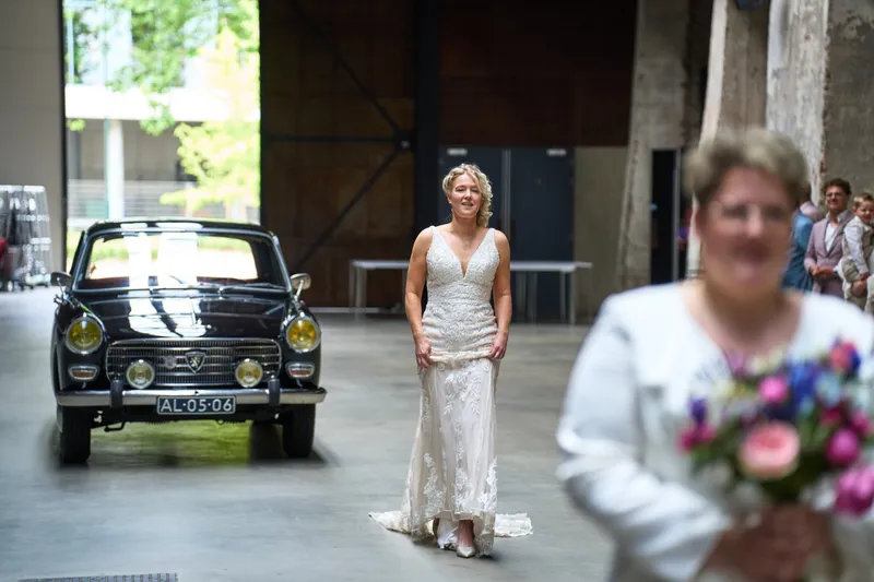 Mireille loopt in haar jurk naar de oldtimer, op de voorgrond een boeket