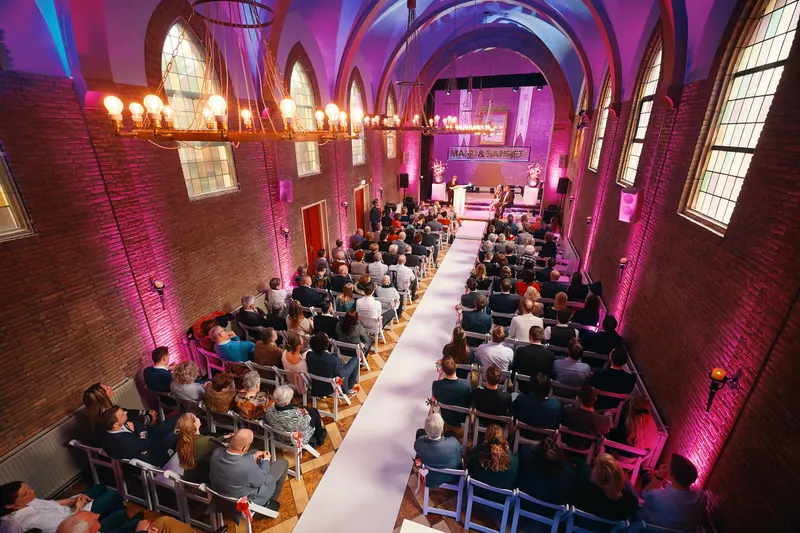 De zaal van De Noviteit met de gasten aan weerszijden van de witte loper, in roze licht