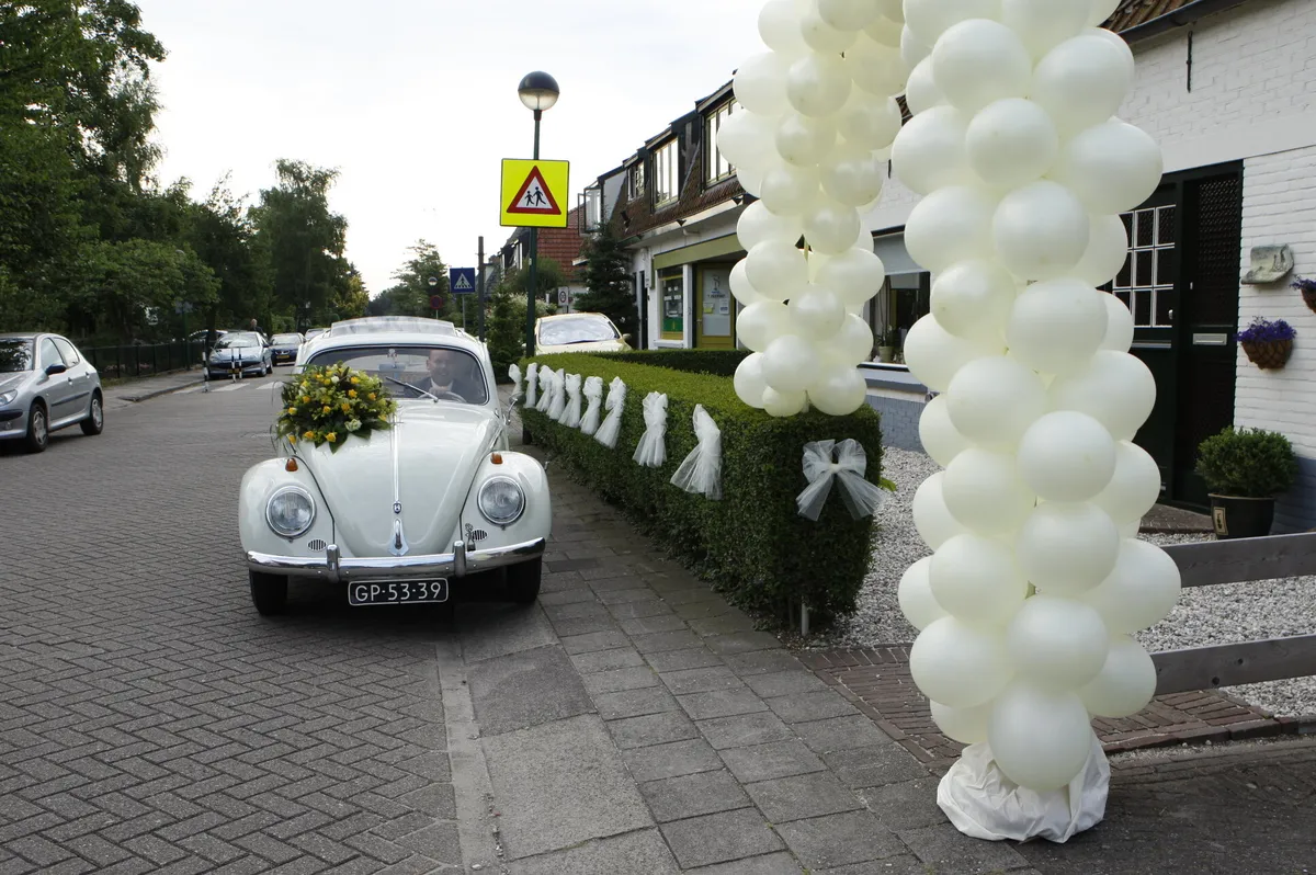 De lichtgrijze Kever voor het huis, met witte ballonnen en strikken langs de heg
