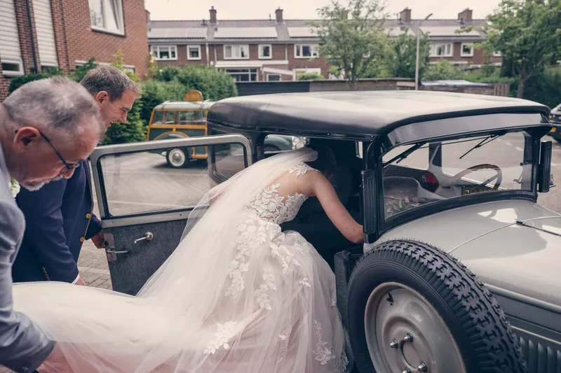 Marjolijn stapt in de oldtimer, iemand helpt met de jurk