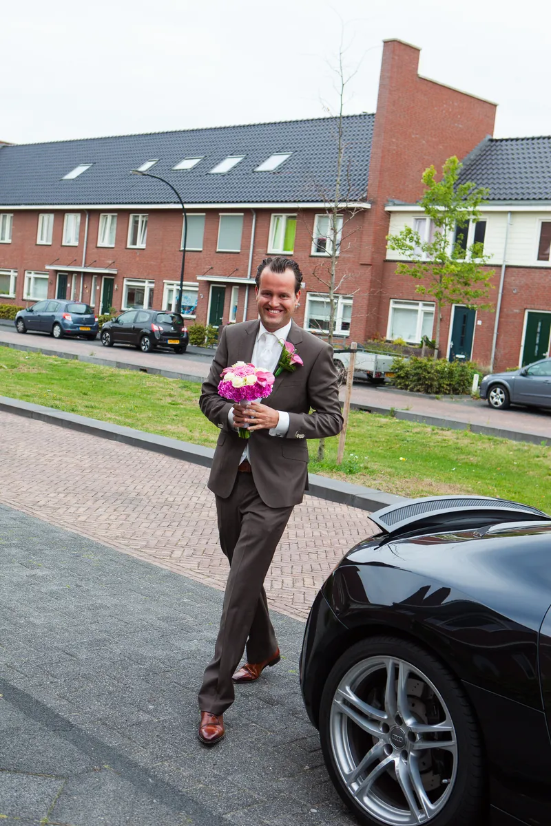 Jan-Willem loopt met het bruidsboeket over straat naar de voordeur, naast de Audi R8