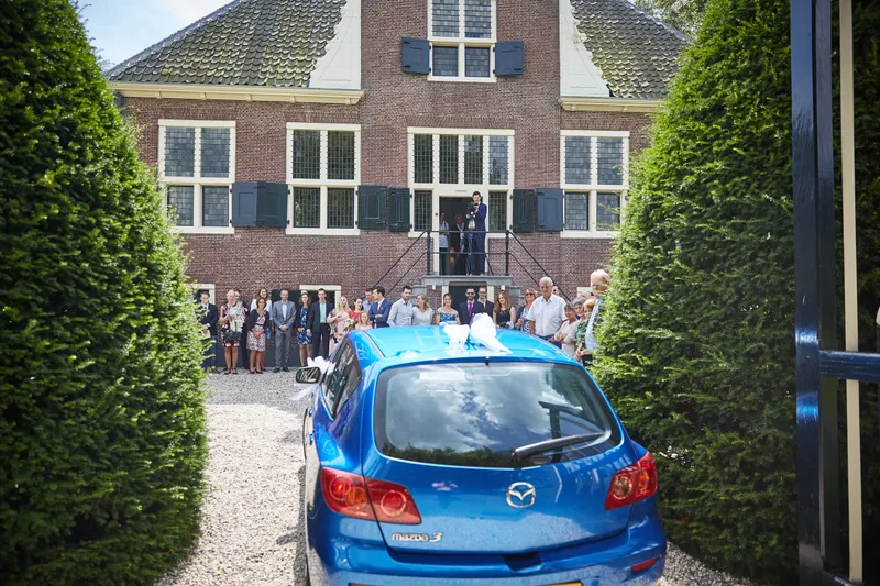 De blauwe trouwauto komt aan bij Hofstede Meerzigt, de gasten staan bij de ingang