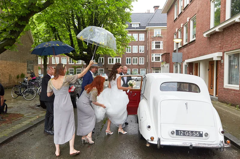 De bruidsmeisjes lopen met paraplu’s naar de Rolls-Royce