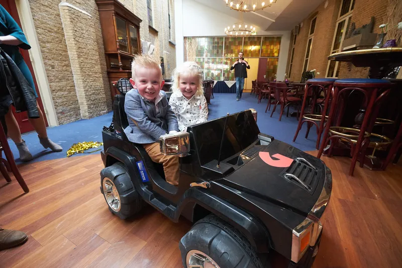 Twee kinderen in een speelgoedjeep tussen de tafels