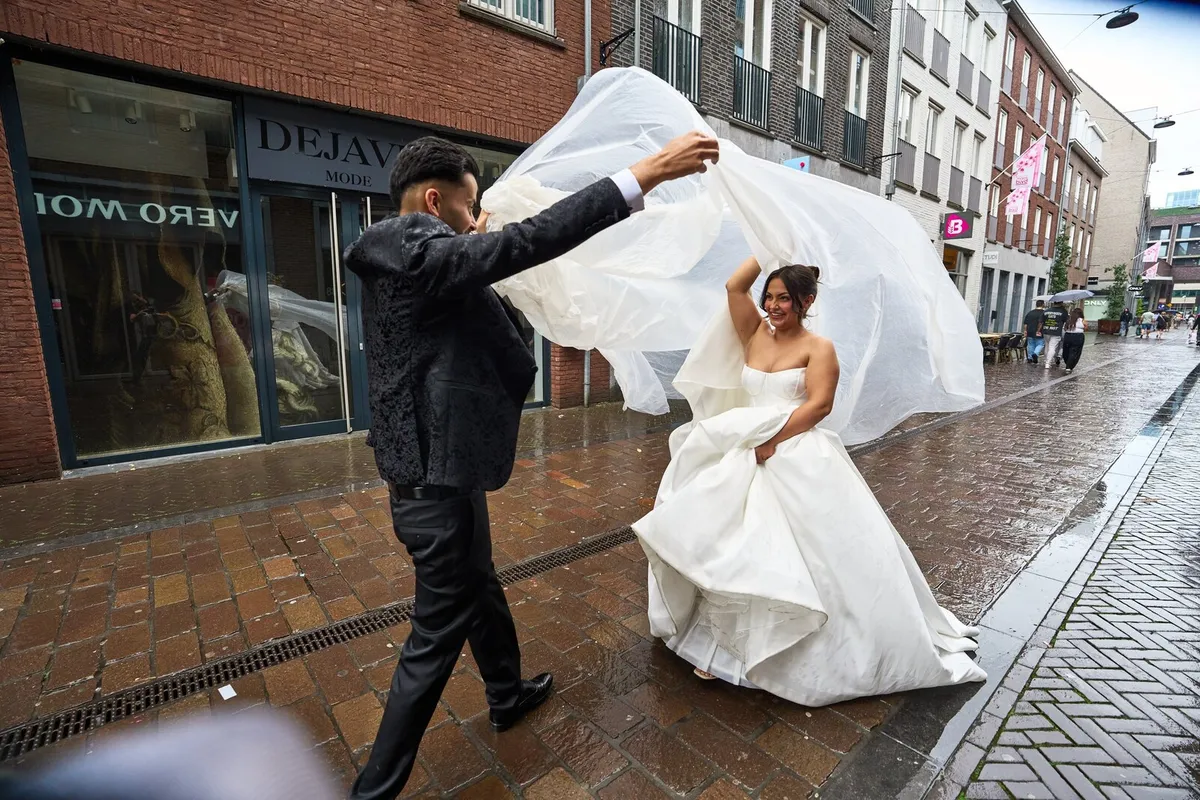 Bruid en bruidegom in een natte winkelstraat, de sluier waait op in de wind