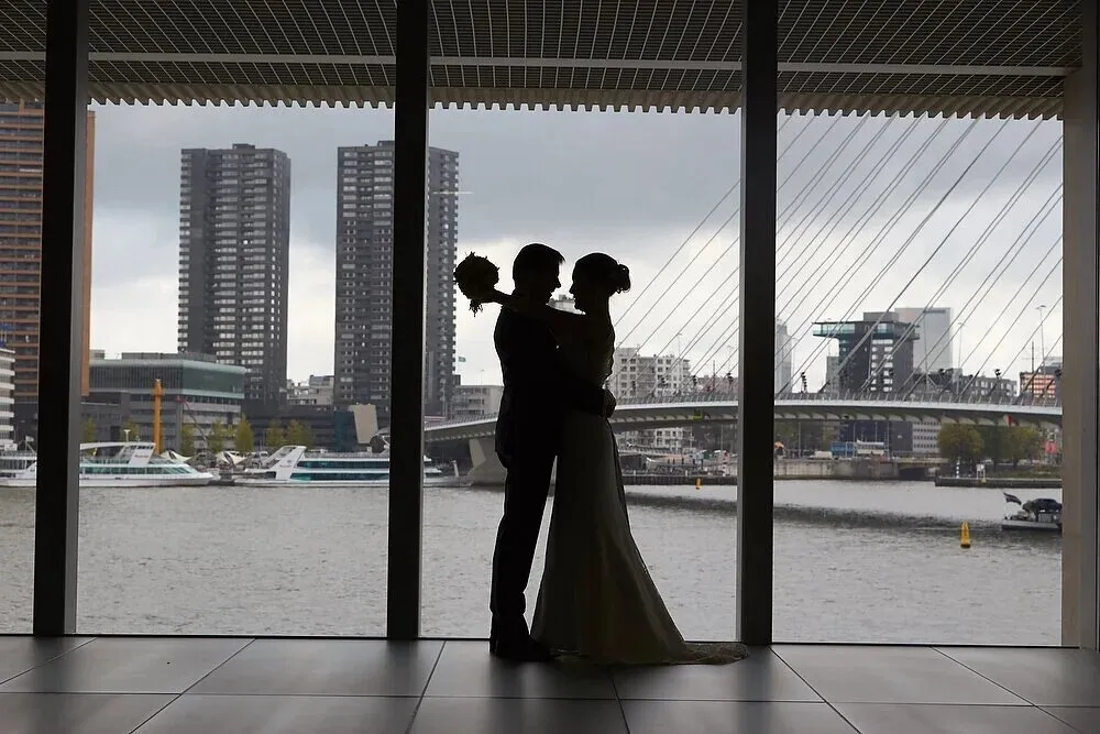 Silhouet van het bruidspaar voor een groot raam, met de Erasmusbrug en de torens van Rotterdam erachter