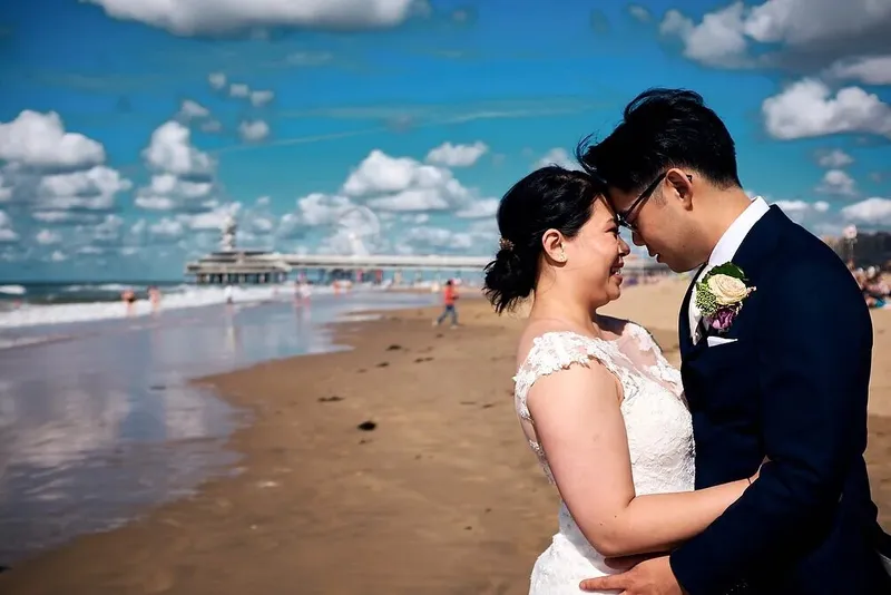 Het bruidspaar hoofd tegen hoofd op het strand van Scheveningen, met de pier erachter