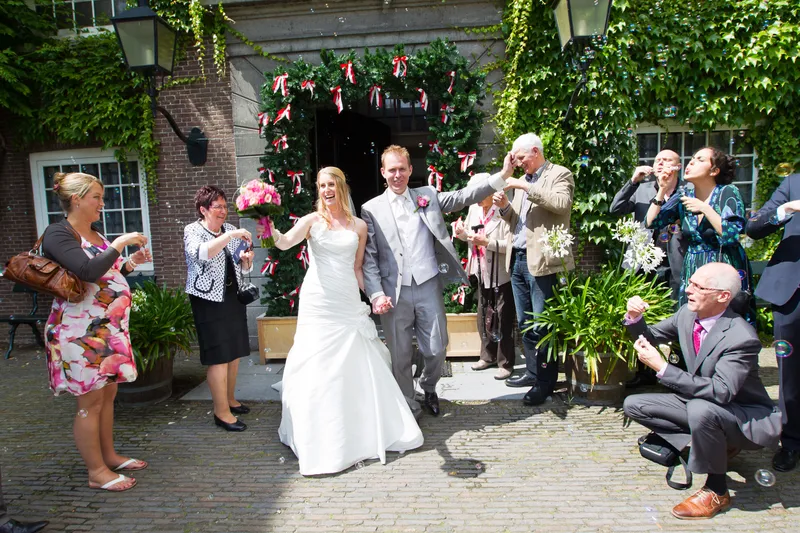 Loes en Jan lopen lachend door de poort met rood-witte linten, terwijl gasten bellen blazen