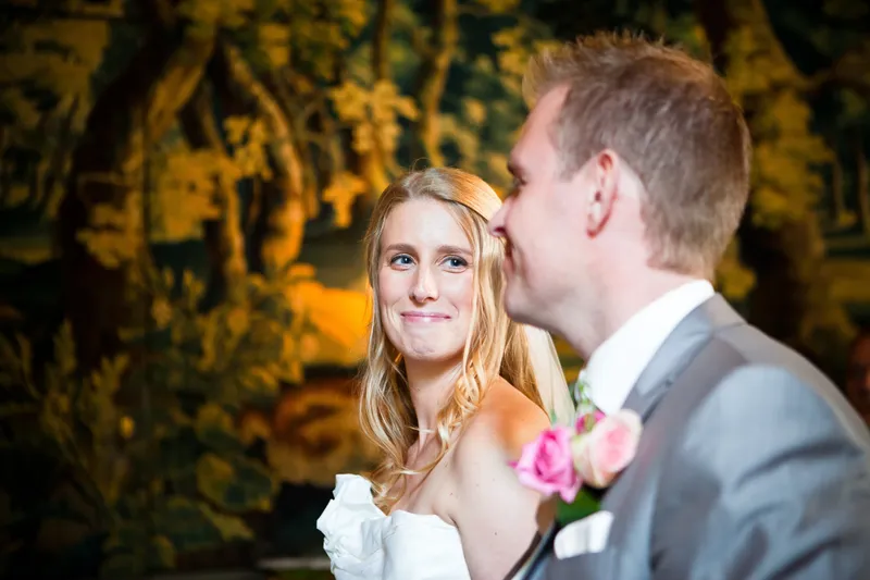 Loes kijkt lachend naar Jan tijdens de ceremonie, met een wandtapijt op de achtergrond