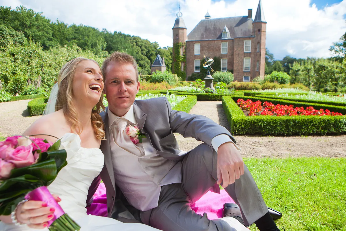 Loes en Jan zitten lachend in het gras, met Slot Zuylen en de tuin achter hen