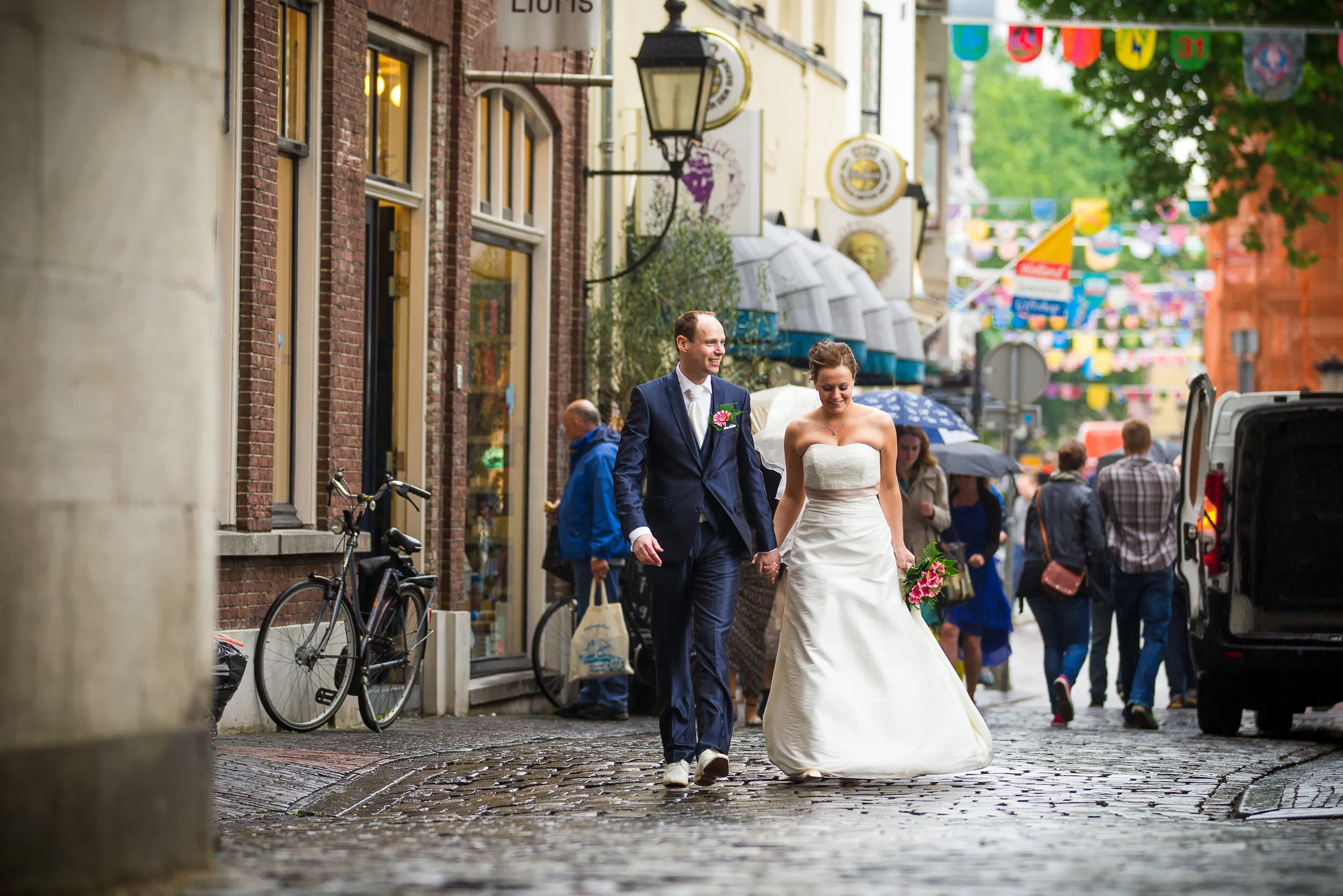 Bruidspaar loopt hand in hand over de natte keien van een winkelstraat in Utrecht, met vlaggetjes boven de straat en winkelend publiek eromheen