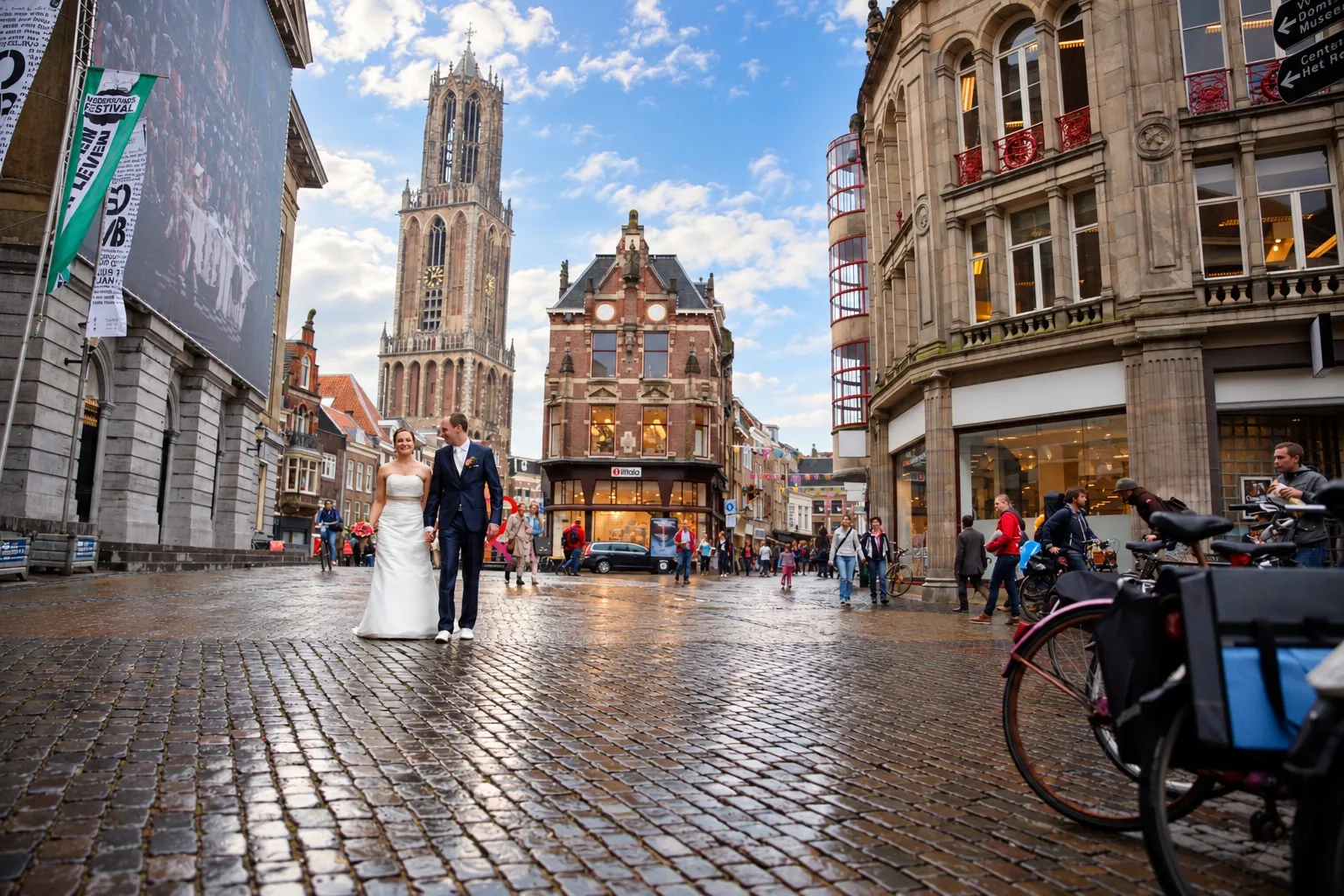 Bruidspaar loopt hand in hand over de natte keien, met de Domtoren achter hen