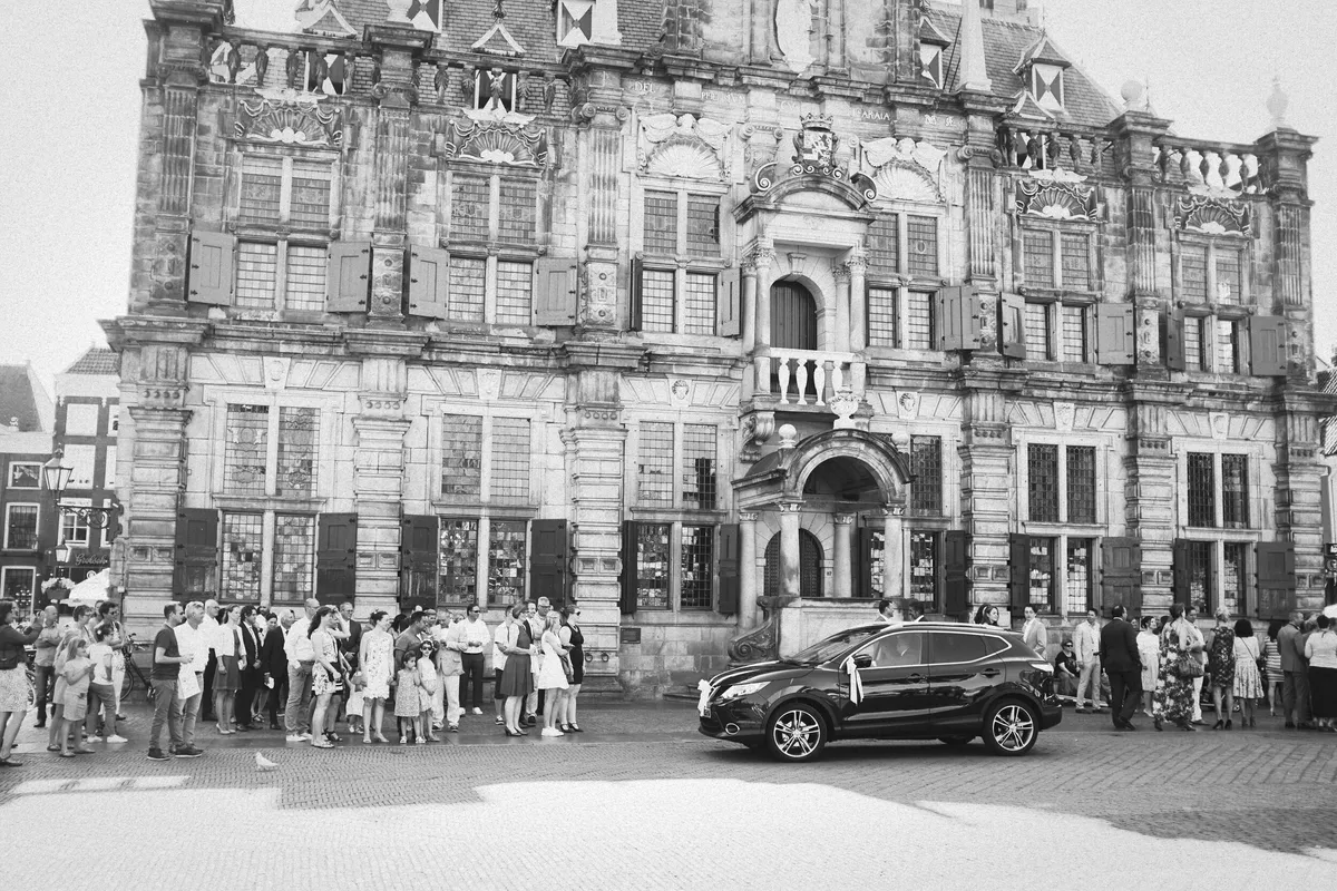Zwart-wit: gasten wachten voor het stadhuis van Delft, met de trouwauto ervoor