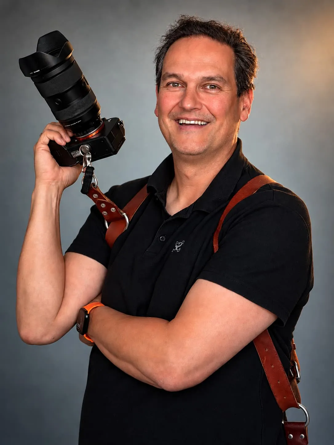 Stefan Segers lacht, met zijn camera omhoog en het leren cameratuig over zijn zwarte polo