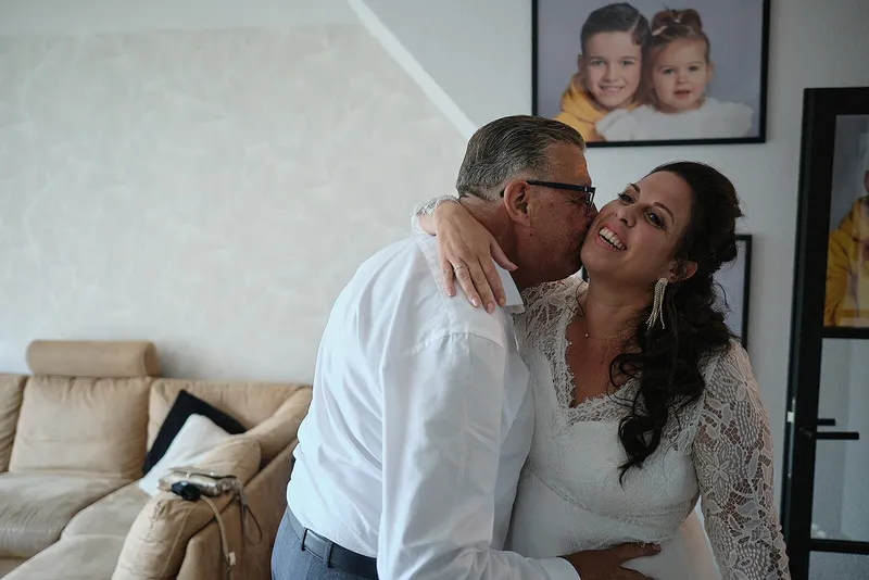 Een man in een wit overhemd omhelst Regina in haar jurk in de woonkamer