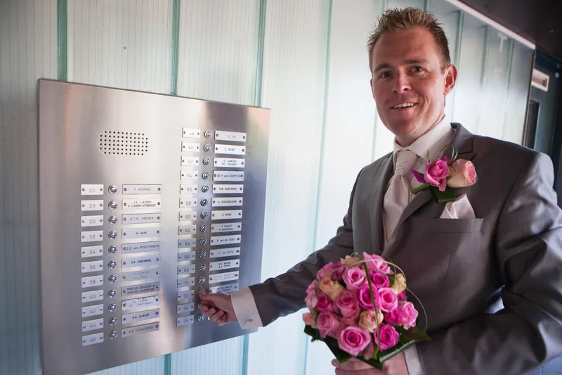 Jan zoekt de juiste bel bij de voordeur, met het roze boeket in zijn hand