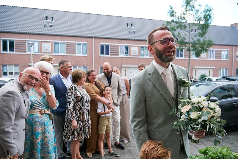 Familie en buren kijken toe terwijl Sander met het boeket bij het huis staat te wachten
