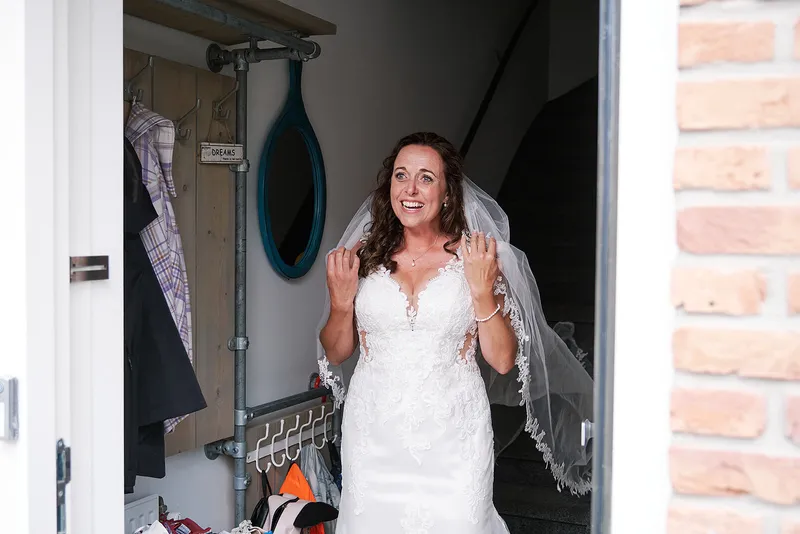 Romana doet de voordeur open in haar trouwjurk, met haar handen voor haar mond
