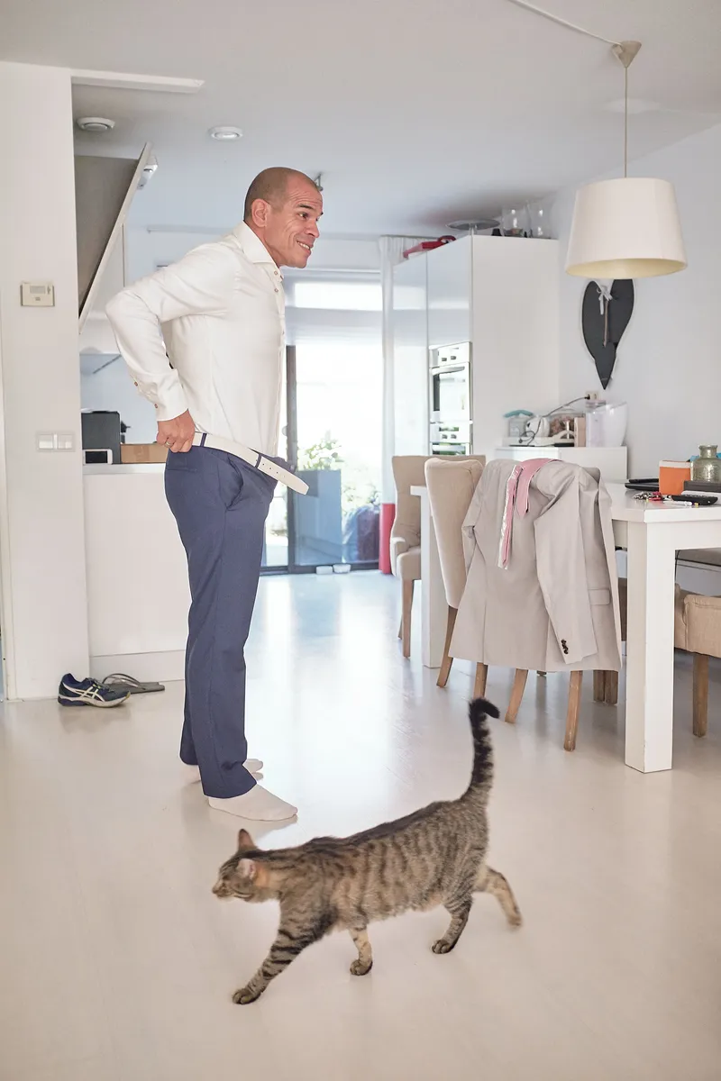 Manuel trekt zijn broek aan in de woonkamer, terwijl de kat voorbijloopt
