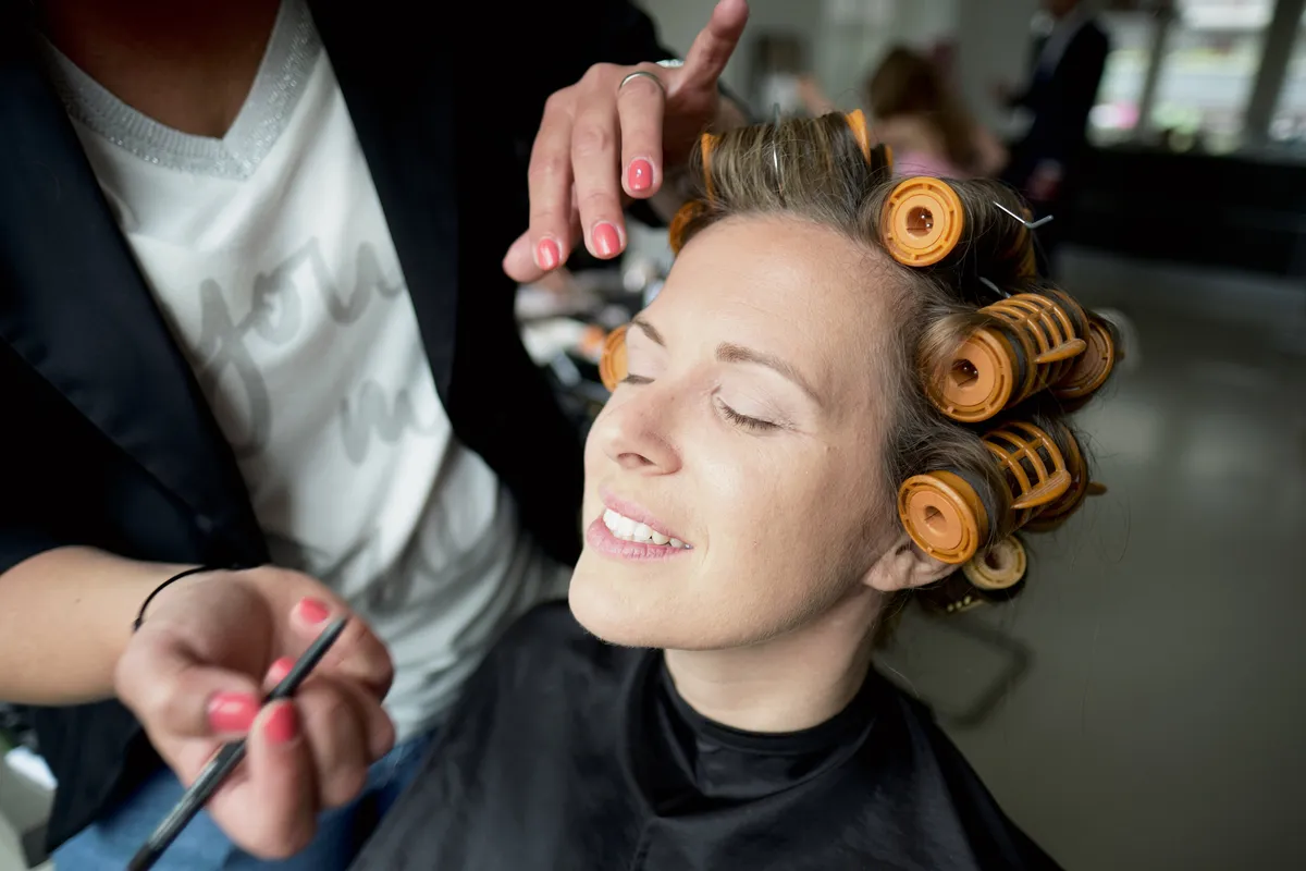 Mary lacht met haar ogen dicht terwijl haar make-up wordt gedaan, met krulspelden in