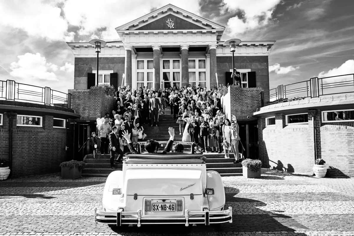 Zwart-wit: de trouwauto en de gasten op de trap van een klassiek gebouw
