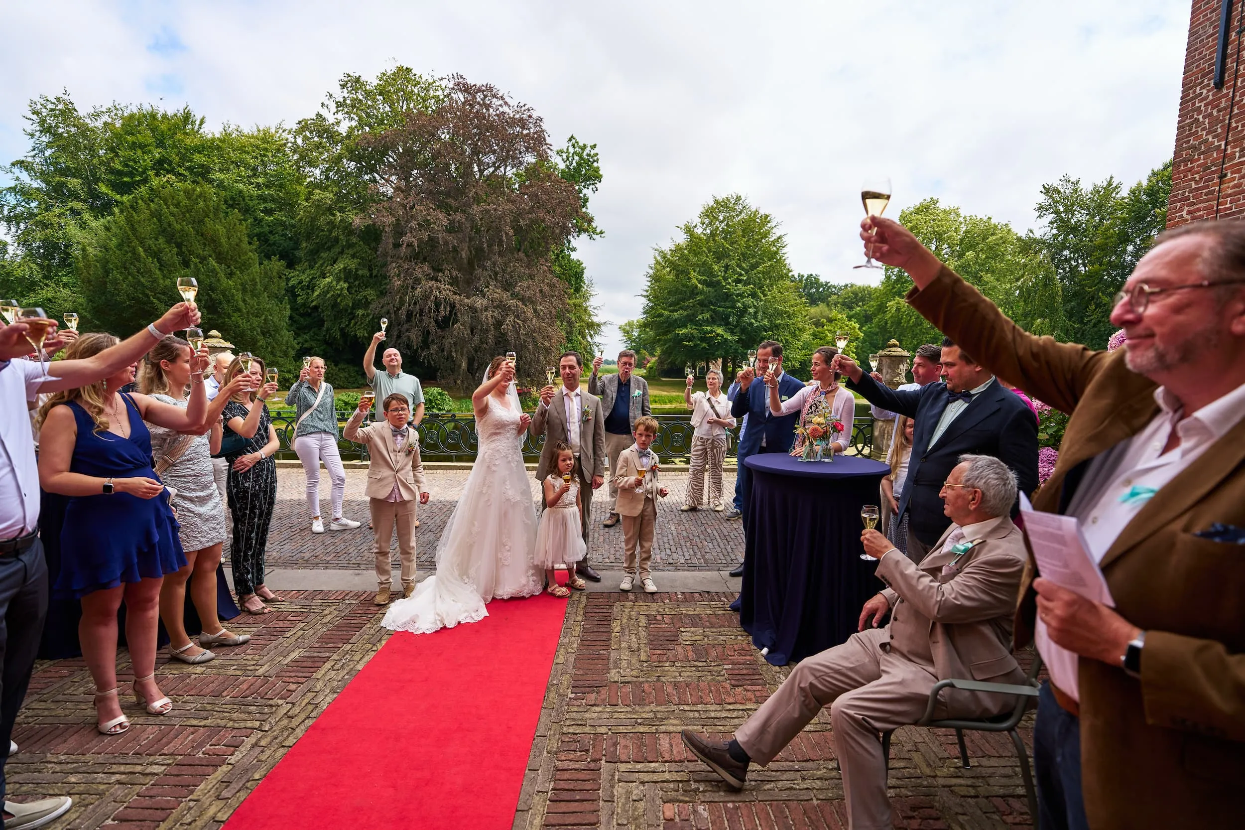 Bruidspaar toost met gasten op rode loper, buiten bruiloft met groene tuin — Trouwshoot Stefan Segers