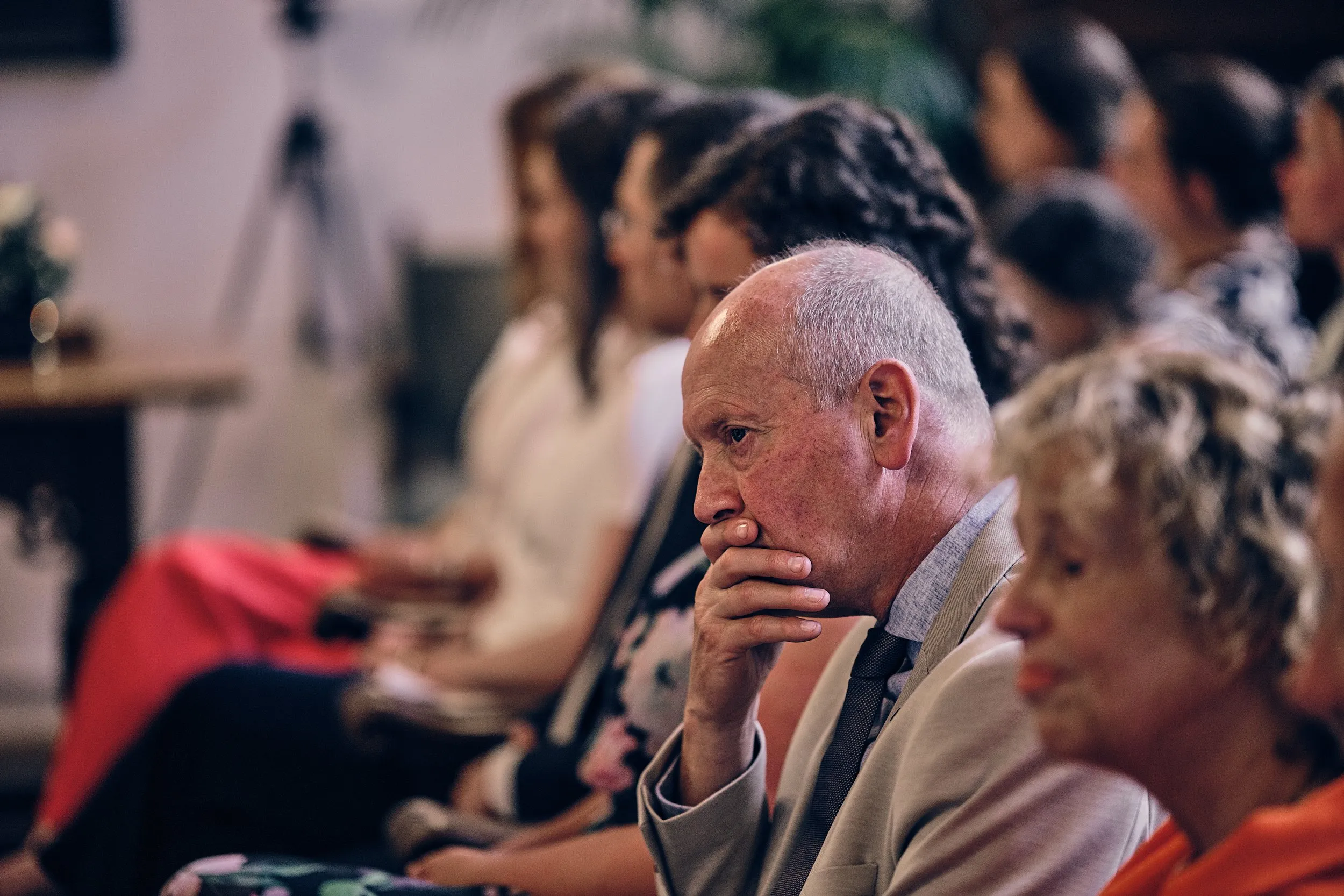 Gastheer in beige pak met donkere stropdas, peinzend kijkend tijdens de ceremonie, omringd door gasten op de bruiloft van Sim