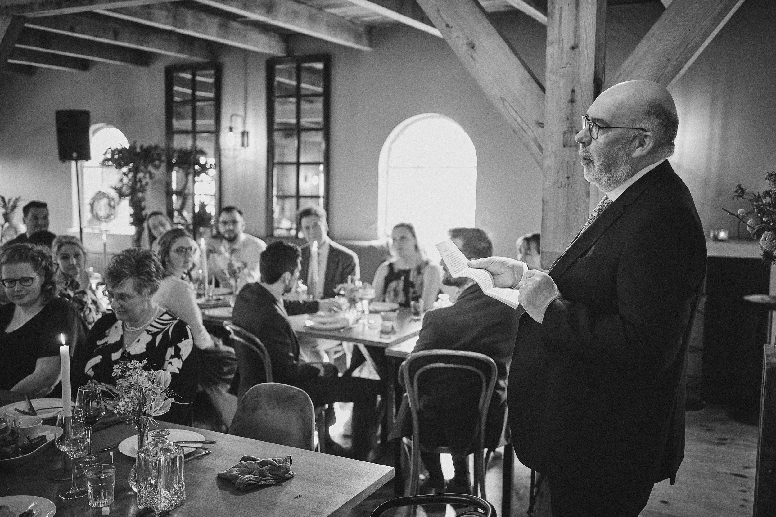 Vader van de bruid houdt speech tijdens het diner op Boerderij Burgersdijk. Gasten luisteren aandachtig in de rustieke zaal m