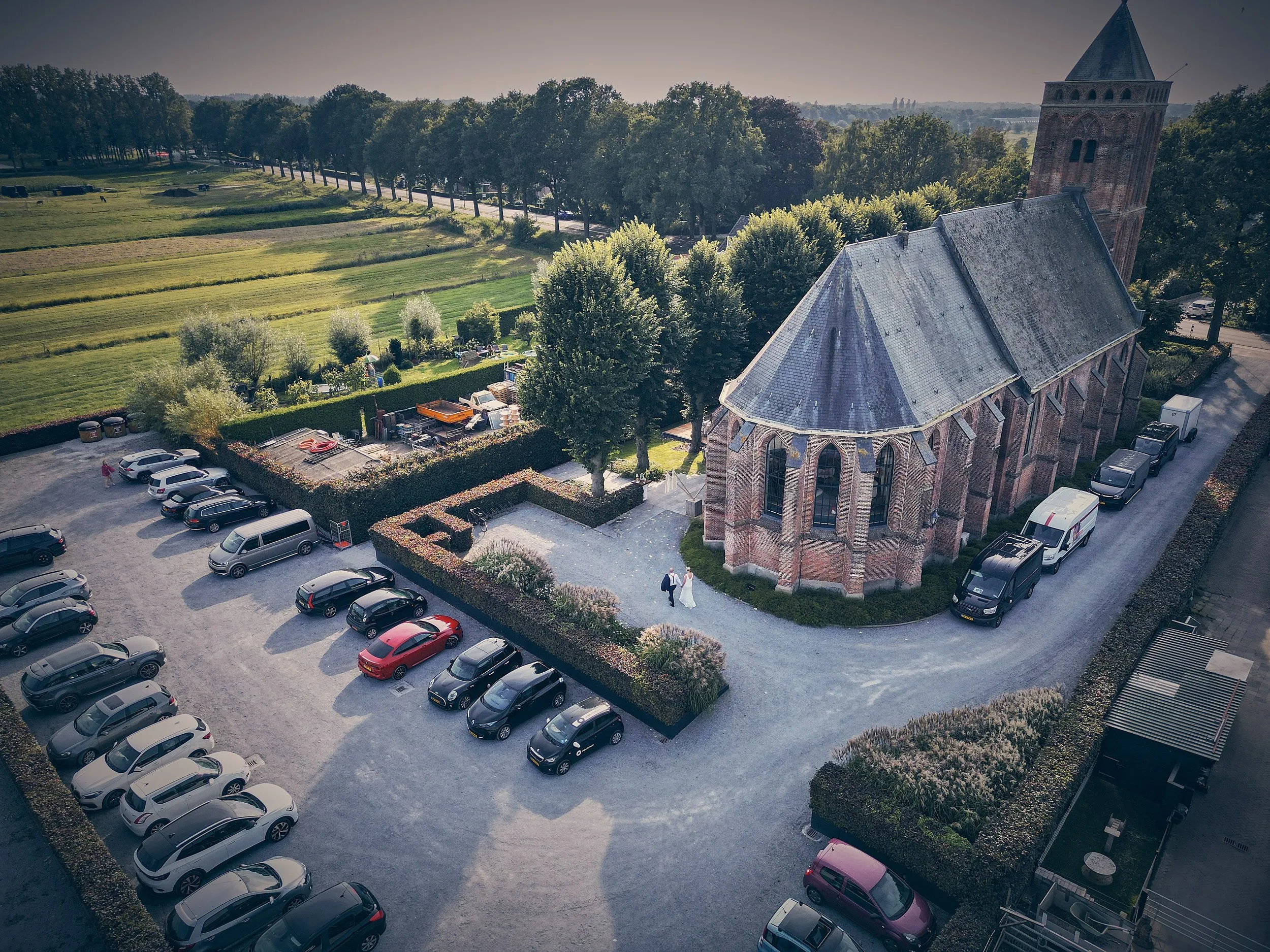 Bruidspaar voor historische kerk met rode dakruiter, omgeven door parkeerterrein en groene landschap