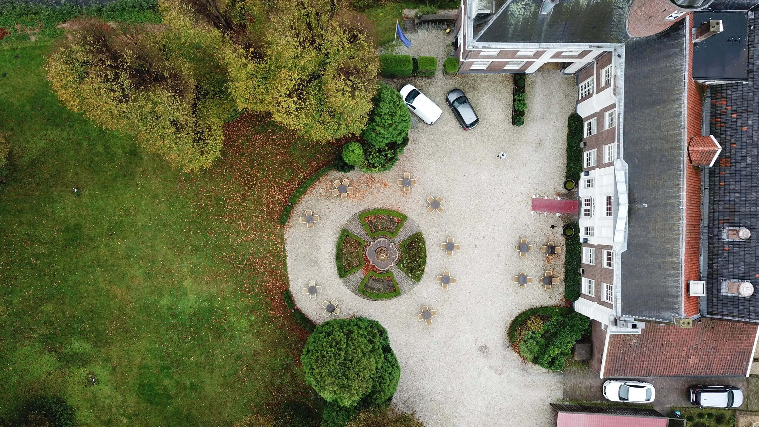 Luchtfoto van een elegant tuinfeest met rond terras, gezellige zithoeken en groene gazon omgeven door herfstkleuren - Trouwsh