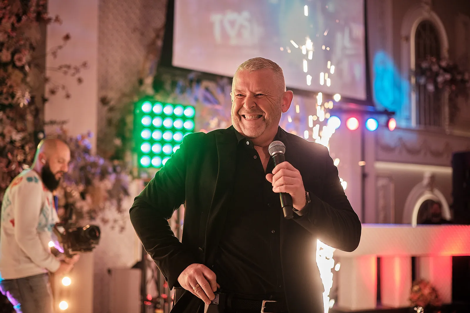Man in zwart blazer spreekt in microfoon tijdens feestelijk bruidsfeest, festive sfeer met gekleurde verlichting