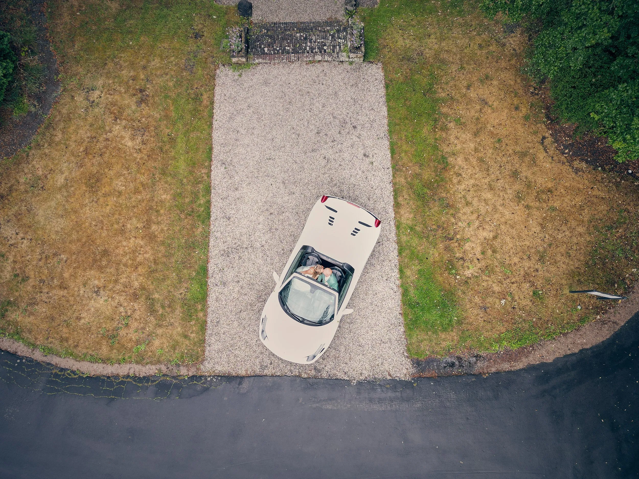 Bruidspaar in witte cabriolet gezien vanuit een drone, rijdend over een landelijke oprijlaan omgeven door groen