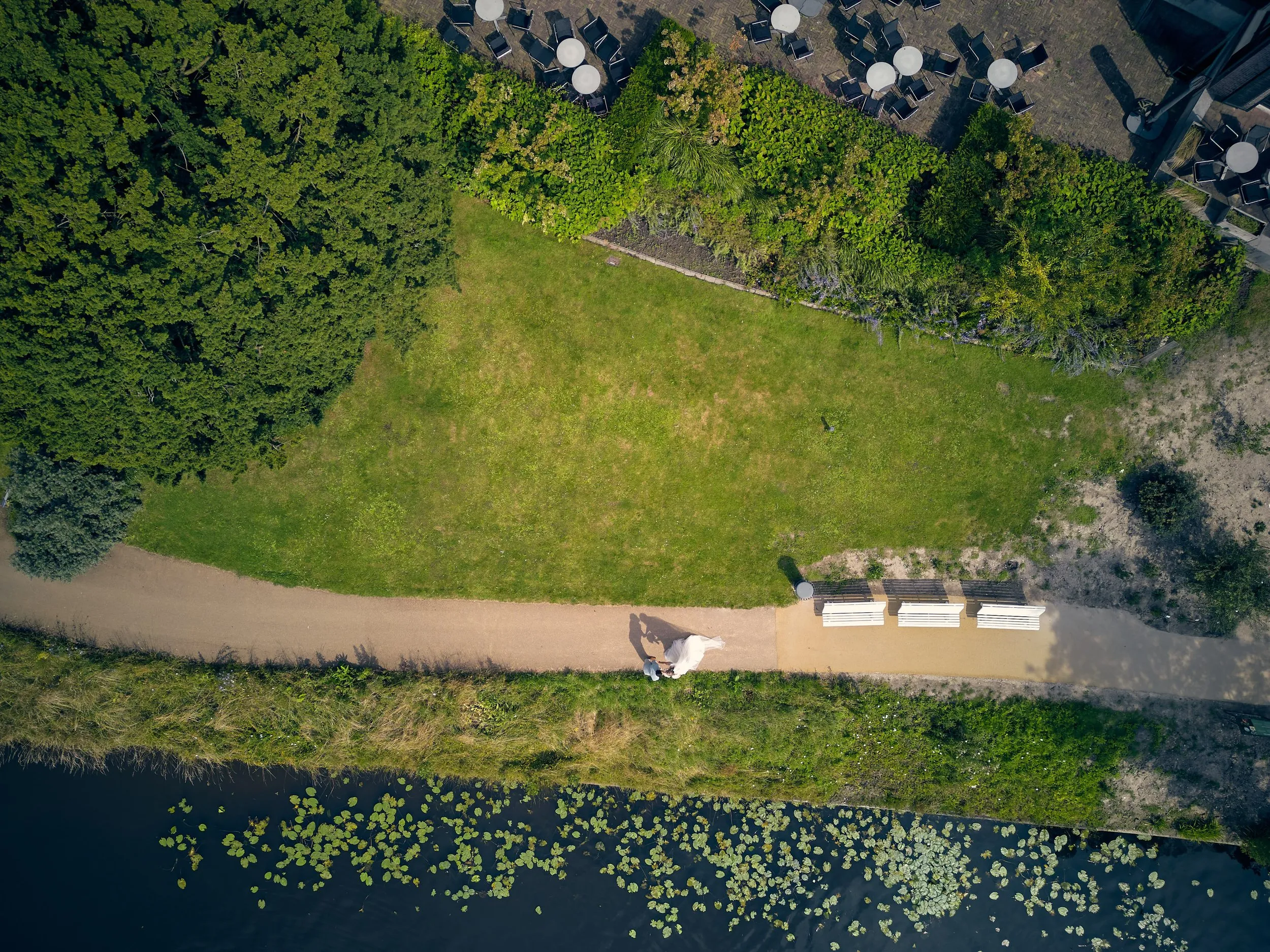 Luchtfoto: bruidspaar Julia en Rens wandelt langs het water bij 't Wapen van Zoetermeer, omgeven door groen en bloeiende wate