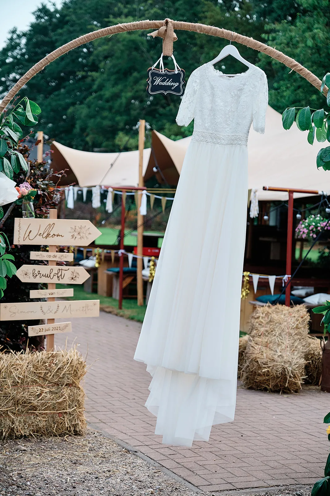 Witte trouwjurk van bruid Mariëth hangt aan houten boog met 'Wedding' bordje, rustieke tuinbruiloft
