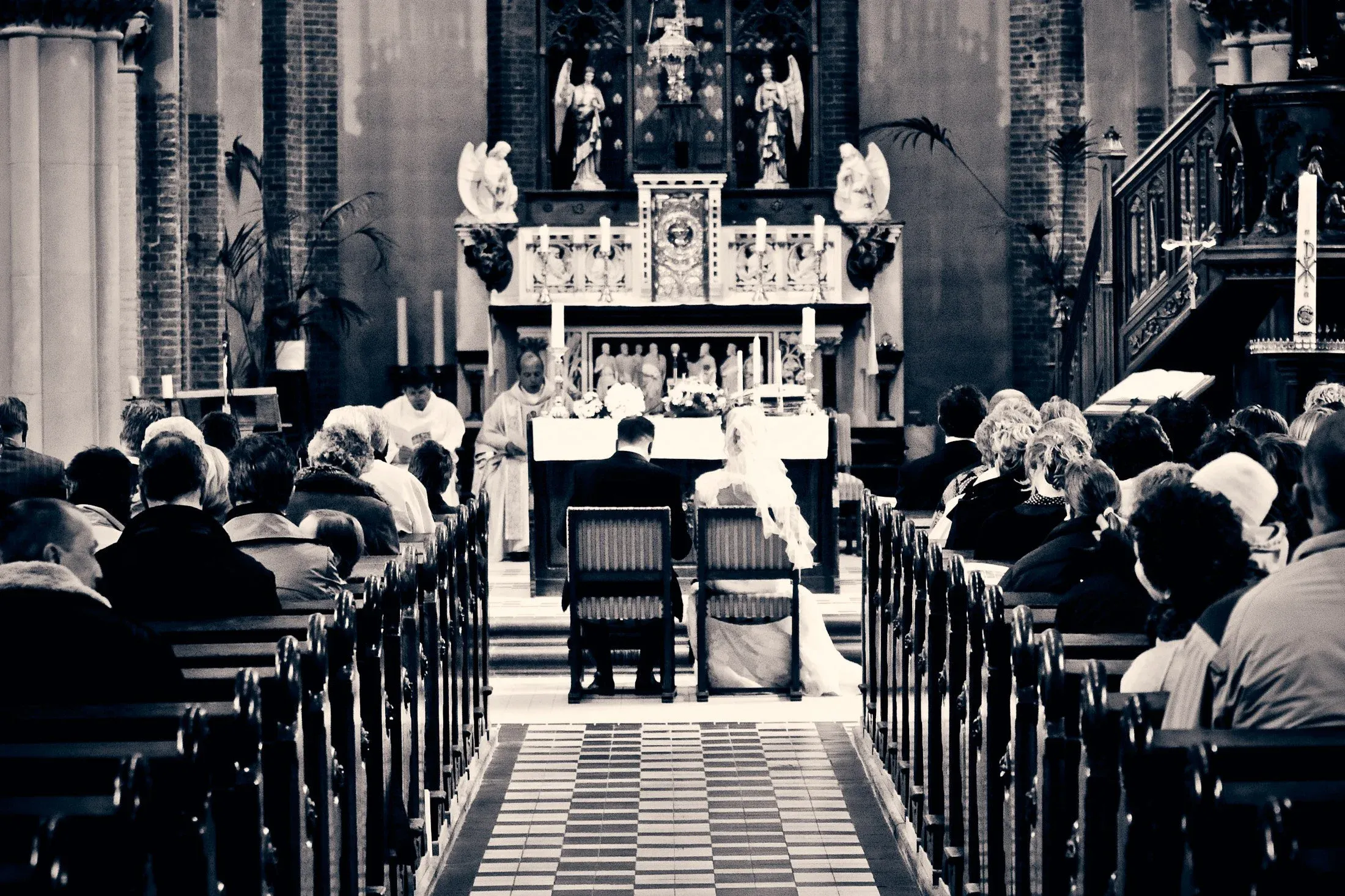 Een huwelijksceremonie in een kerk met veel mensen die aan weerszijden van de kerkbanken zitten, het bruidspaar zit vooraan bij de altaar.