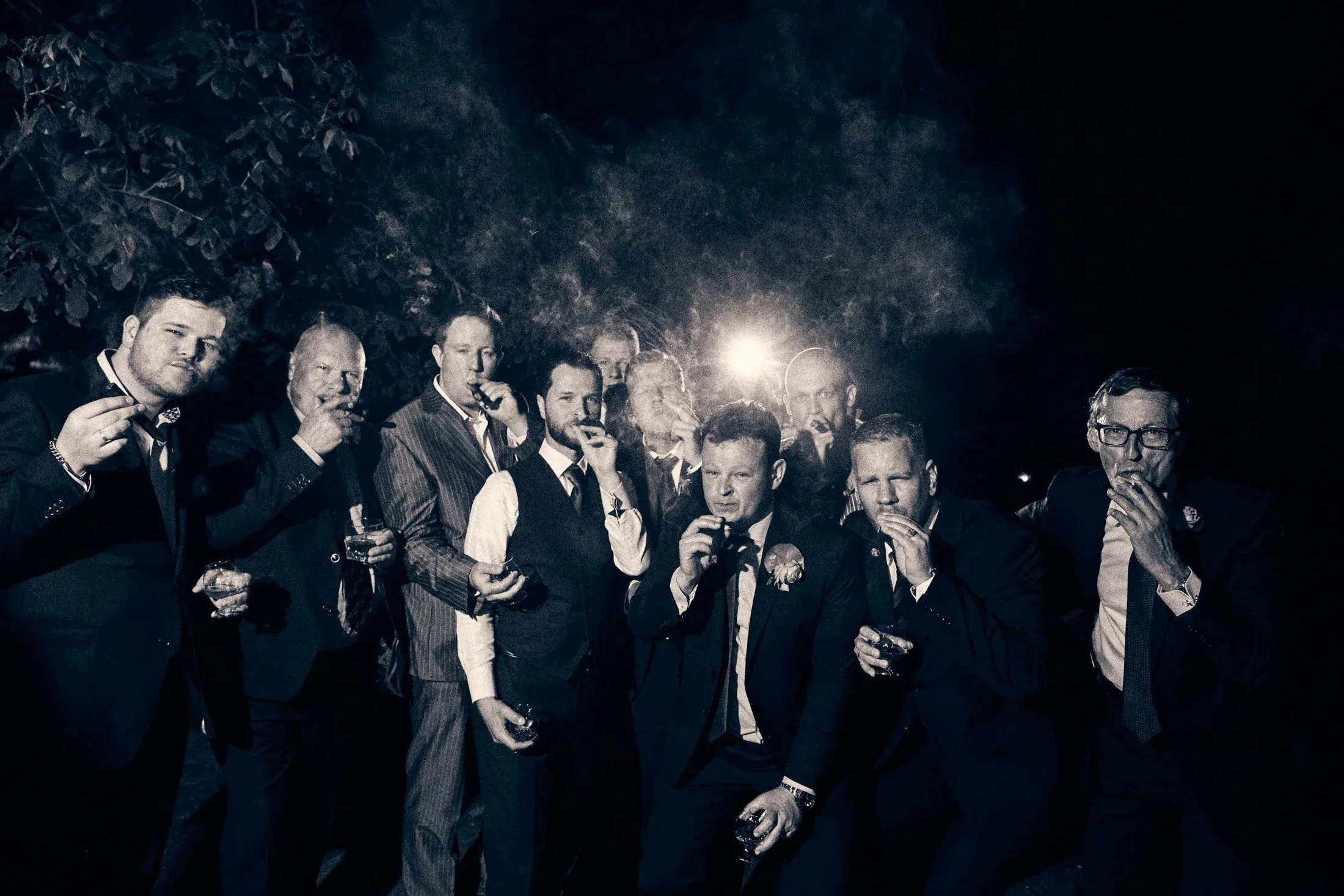 Een groep mannen in formele kleding die samen wijn drinken tijdens een nachtelijke sociale gebeurtenis, met een donkere achtergrond en een lichtbron op de achtergrond.