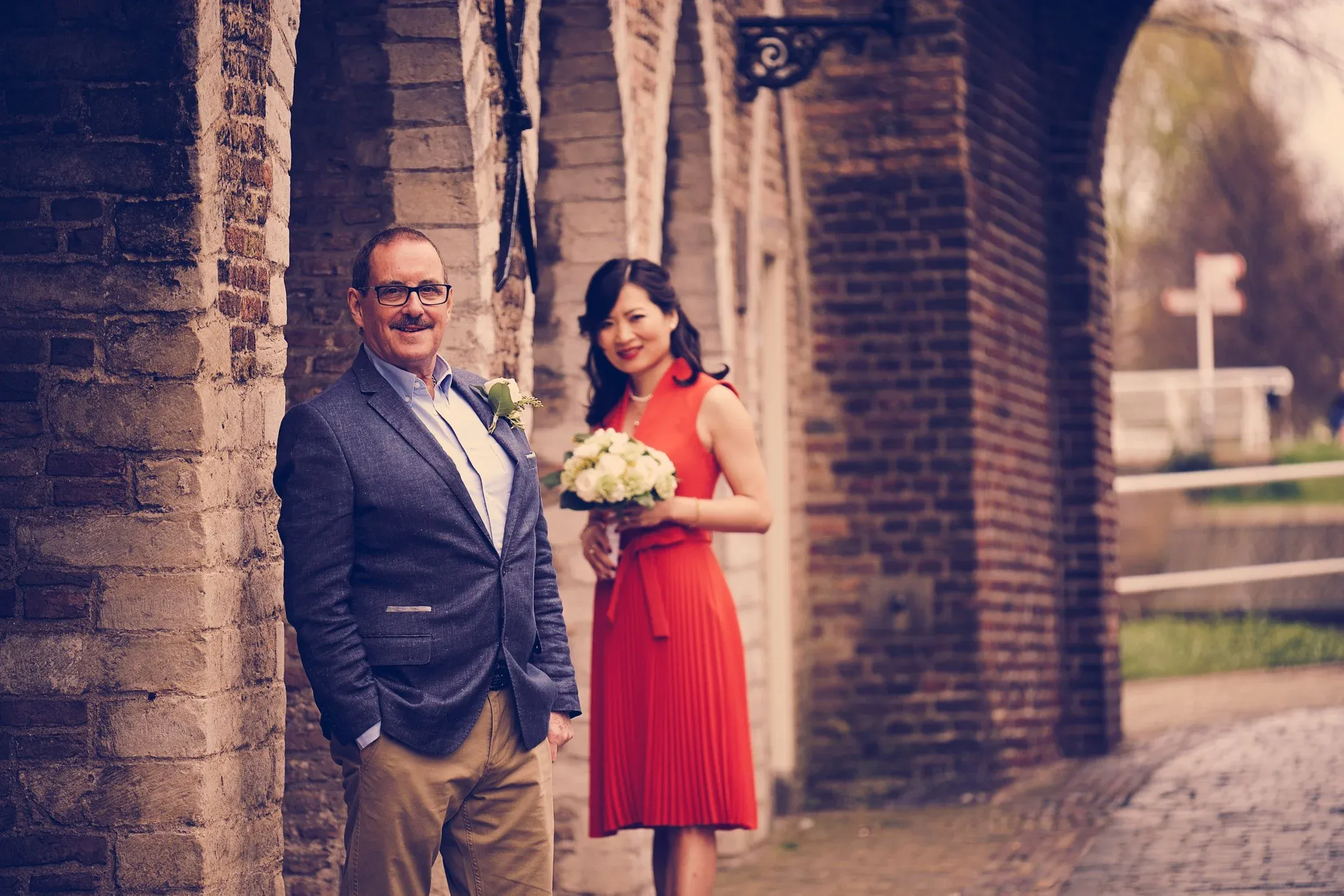 Een man in een grijs jasje en een vrouw in een rode jurk staan naast een oude stenen muur, mogelijk bij een bruiloft, met een bloemenboeket in haar handen.