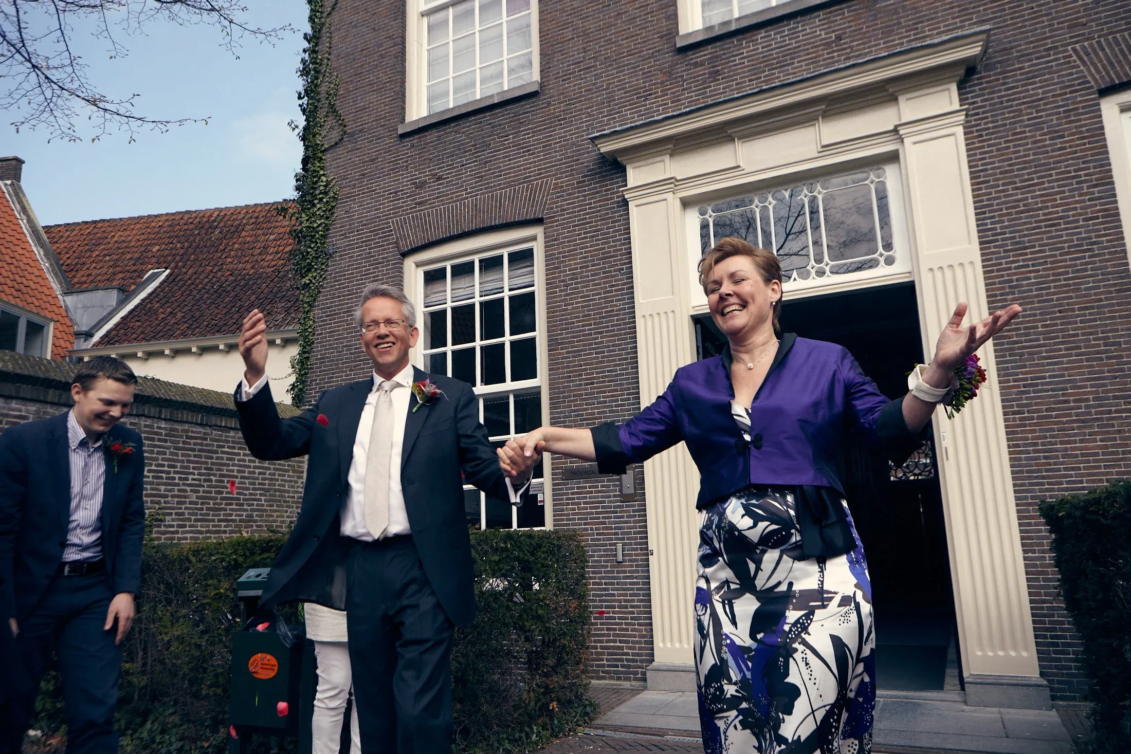 Vrouw en man die elkaar de hand schudden voor een kerk, lachend en feestelijk gekleed, terwijl een jongere man toekijkt. Ze vieren waarschijnlijk een bruiloft buiten voor een bakstenen gebouw.