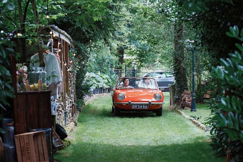 Een oranje oldtimer rijdt door de groene tuinlaan naar het huis