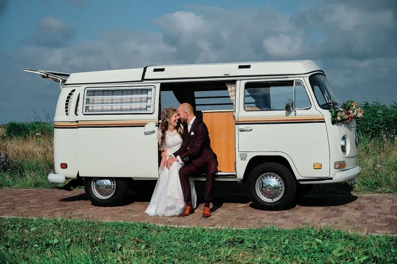 Bruidspaar zit in de zijdeur van een witte VW T2-camper met bloemen op de voorkant