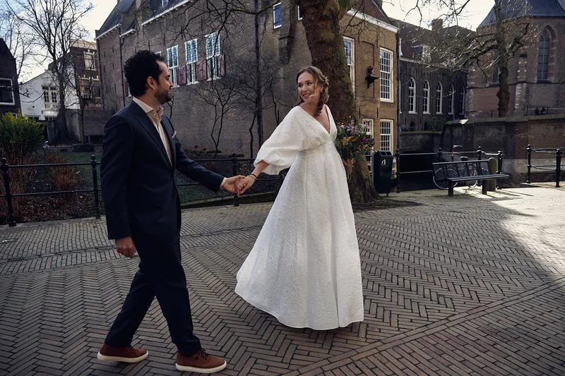 Anouk en Raphaël lopen hand in hand over de klinkers, haar jurk waait mee