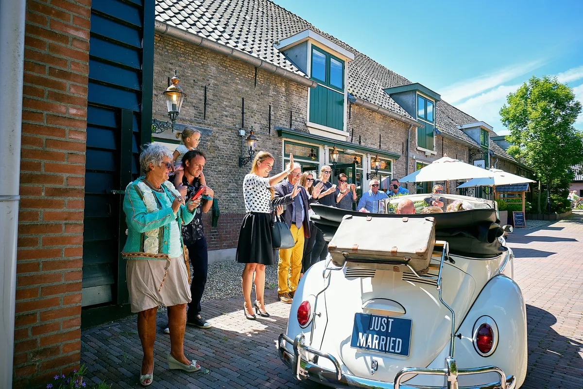Gasten zwaaien bij de witte Kever met Just Married-bord voor ’t Wapen van Zoetermeer