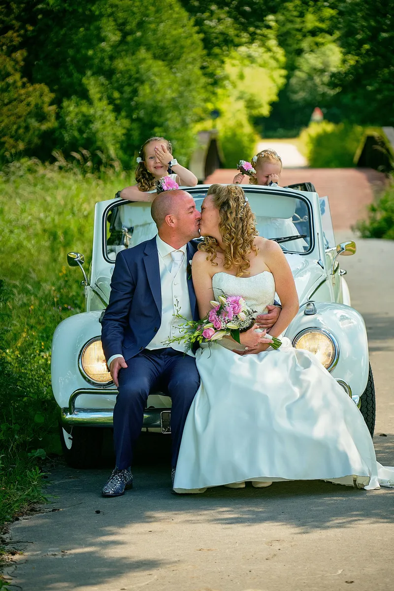 Chantal en Ramon kussen op de motorkap van de witte Kever, de kinderen kijken over de stoel