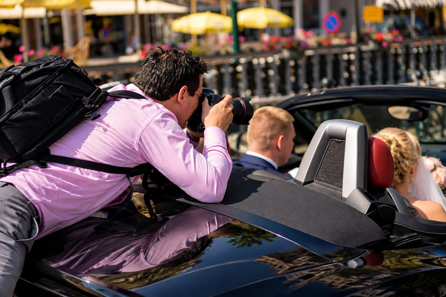 Stefan hangt met zijn camera over een cabrio om het bruidspaar op de achterbank te fotograferen