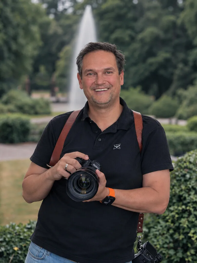 Stefan Segers lachend met zijn camera in een park, met een fontein achter hem