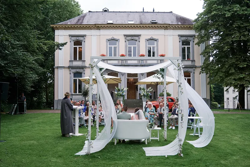 De ceremonieplek op het gras voor het witte landhuis, met witte doeken en bloemen